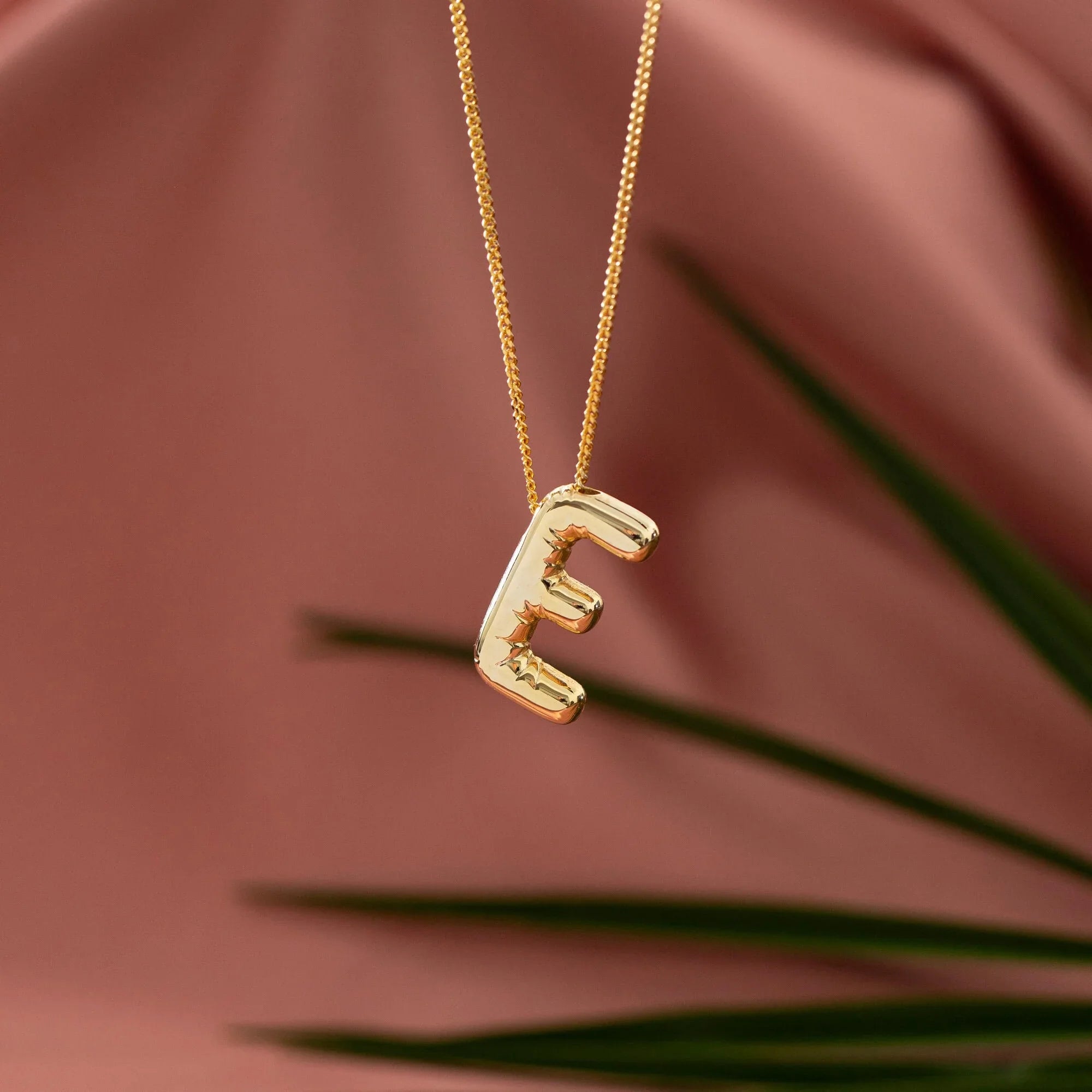 Gold initial E pendant necklace on a pink satin background with green leaf accent.