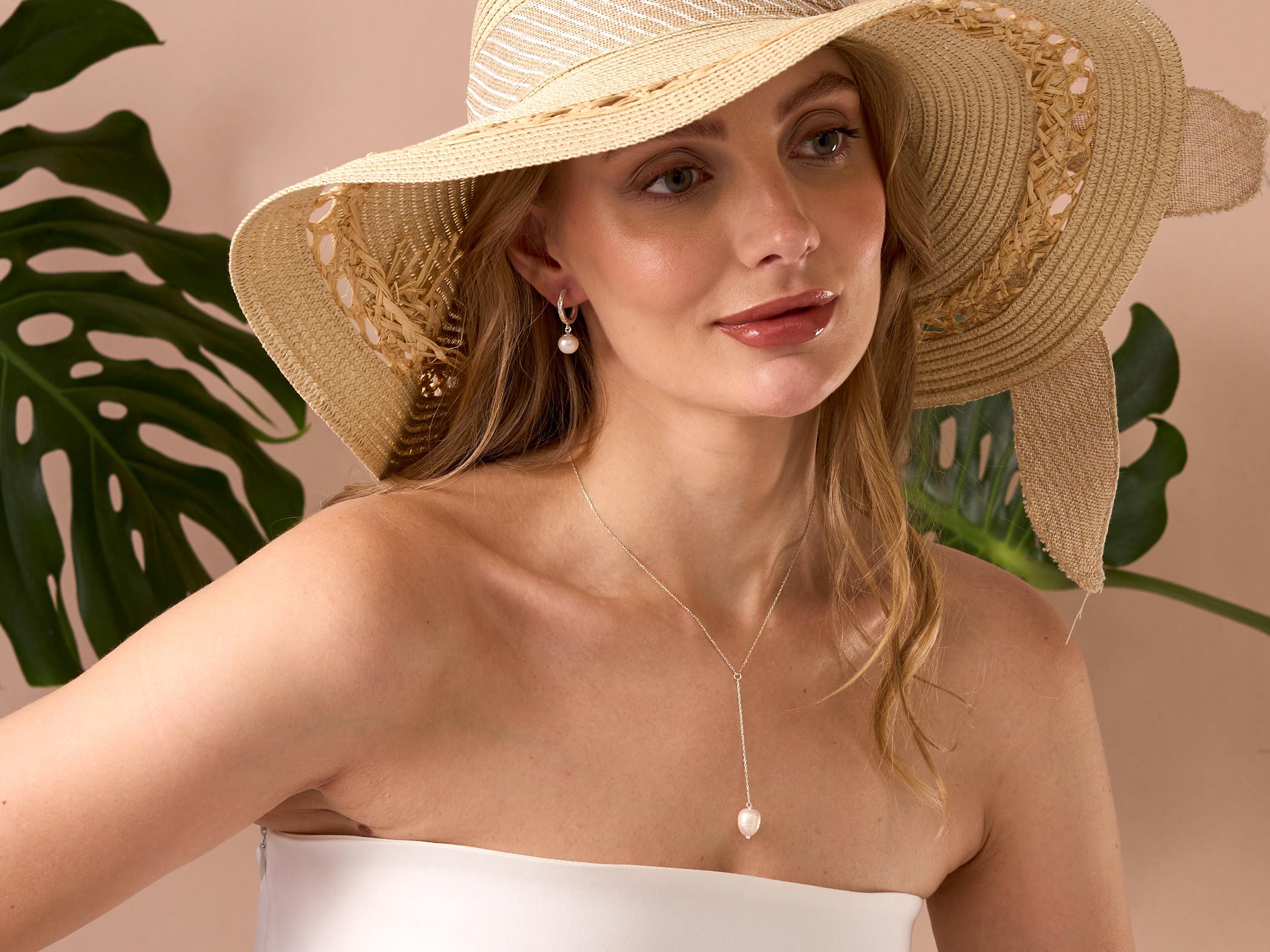 Woman wearing a straw hat and white top with pearl a necklace, surrounded by green leaves.